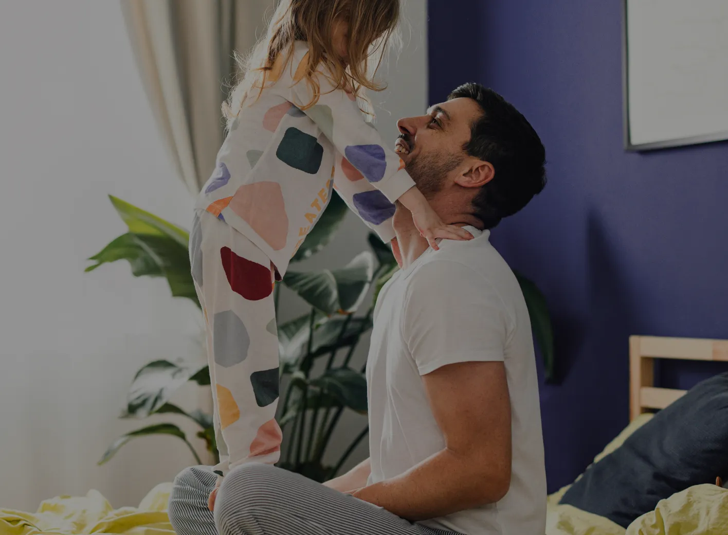 A man kneels on a bed, smiling at a young girl, his daughter, standing in front of him. The girl is in colorful pajamas and rests her hands on his shoulders. The room has a purple wall, large green plants, and a window with curtains.