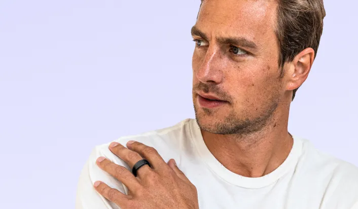 A man with short hair wearing a white t-shirt looks to the side with a thoughtful expression. He rests one hand on his shoulder, wearing a black ring. The background is a light purple shade.